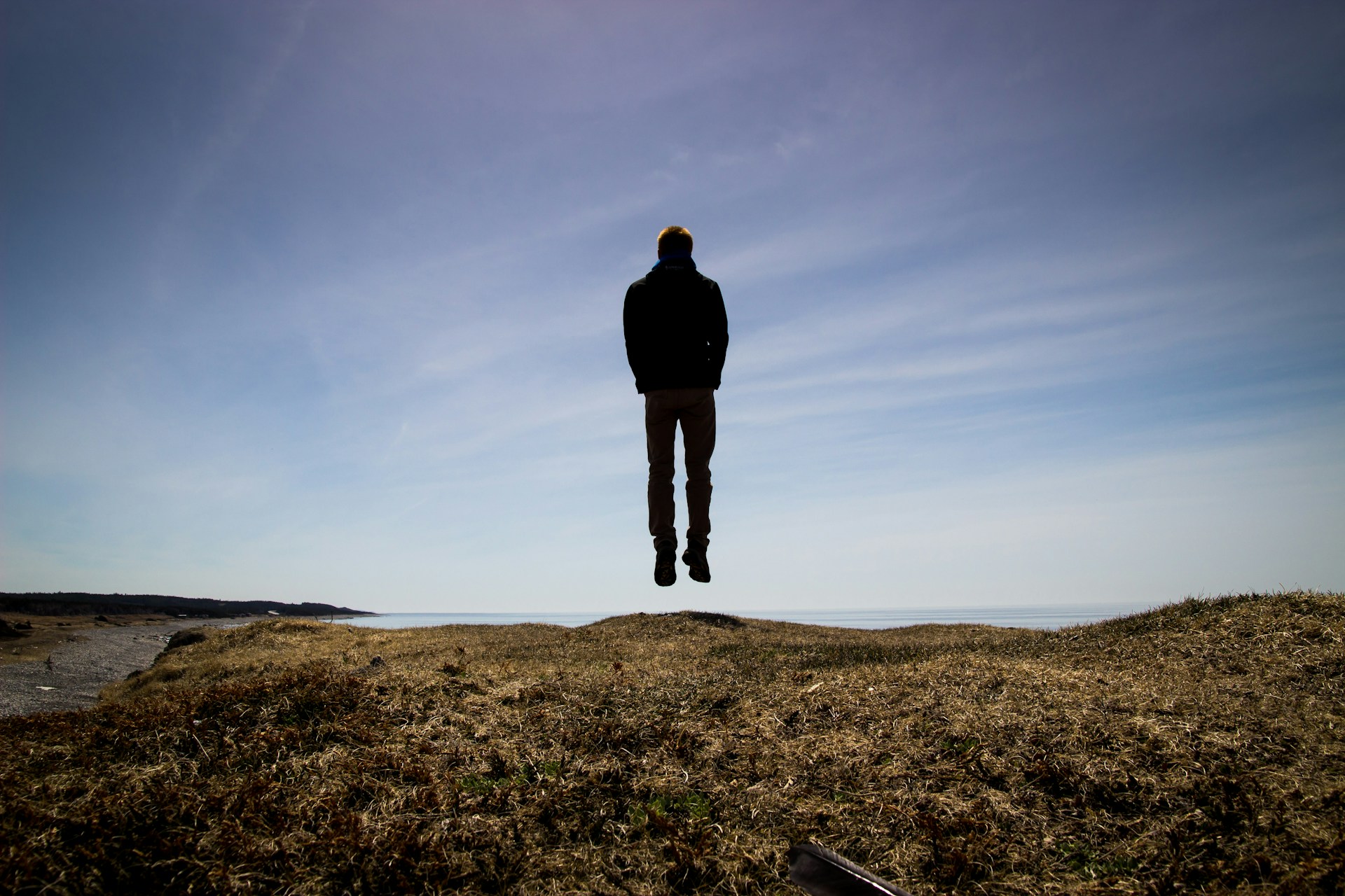 Guy jumping straight up from behind on a grassy knoll. Sky in background.